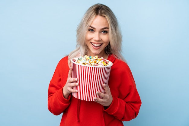 Teenager girl over isolated blue background holding a big bucket of popcorns