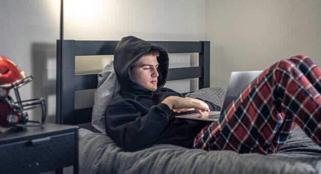 A teenager guy sits in a room on a bed and uses a laptop