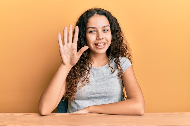 Free photo teenager hispanic girl wearing casual clothes sitting on the table showing and pointing up with fingers number five while smiling confident and happy.