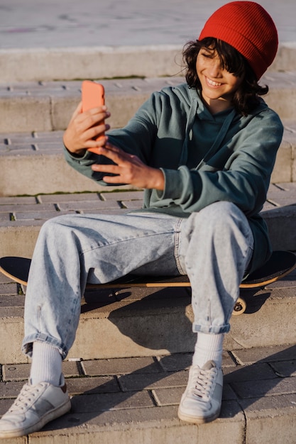Foto gratuita adolescente all'aperto seduto su skateboard e prendendo selfie