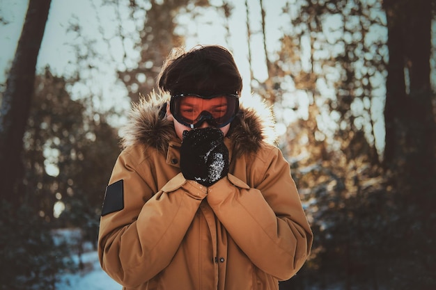 Teenager in protective goggles is warming up his hands at winter forest.