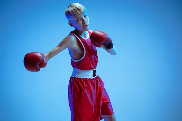 Teenager in sportswear boxing isolated on blue studio background in neon light. Novice male caucasian boxer training hard and working out, kicking. Sport, healthy lifestyle, movement concept.