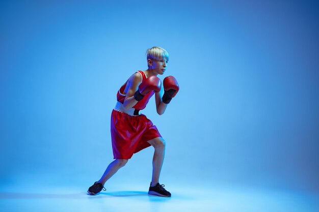 Teenager in sportswear boxing isolated on blue studio background in neon light. Novice male caucasian boxer training hard and working out, kicking. Sport, healthy lifestyle, movement concept.