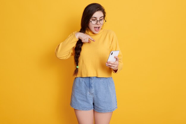 Teenager with opened mouth pointing at smart phone's screen which holding in hand