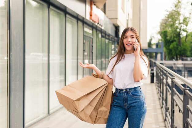 Telling friend about sales. Beautiful young smiling woman holding shopping bags and talking on the mobile phone while standing outdoors