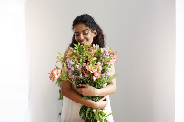 Tender african woman florist smiling holding bouquet of alstroemerias over white wall. Closed eyes.