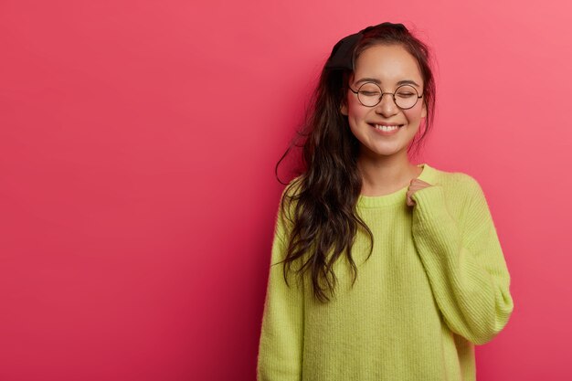 Tender alluring female model grins happily at camera, keeps arm raised, wears green jumper and round transparent glasses, thinks about something pleasant