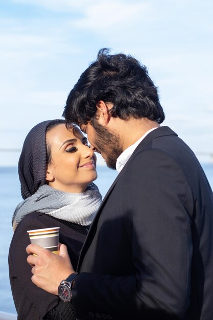 tender arabic couple during walk. woman with covered head and man in suit standing being fond of each other, holding disposable cups of coffee. love, affection concept