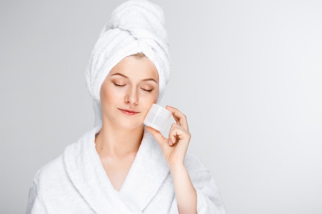 Нежная белокурая женщина с банным полотенцем на волосах показывает крем