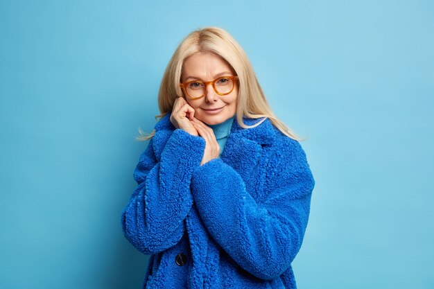 Tender blonde European woman keeps hands near face looks pleasantly has self confident expression wears spectacles fashionable winter fur coat.