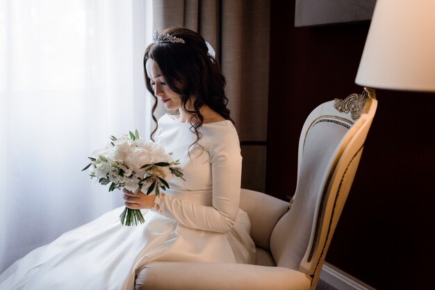 Tender brunette bride is sitting on the armchair, dressed in diadem and holding wedding bouquet made of white eustomas and peonies