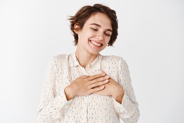 Tender brunette girl in blouse dreaming about something romantic close eyes and smile with hands on heart standing over white background