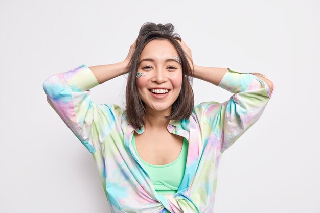 Tender cheerful Asian girl has healthy skin looks with joy  keeps hands on head smiles broadly being in good mood enjoys free time dressed in colorful shirt poses against white wall