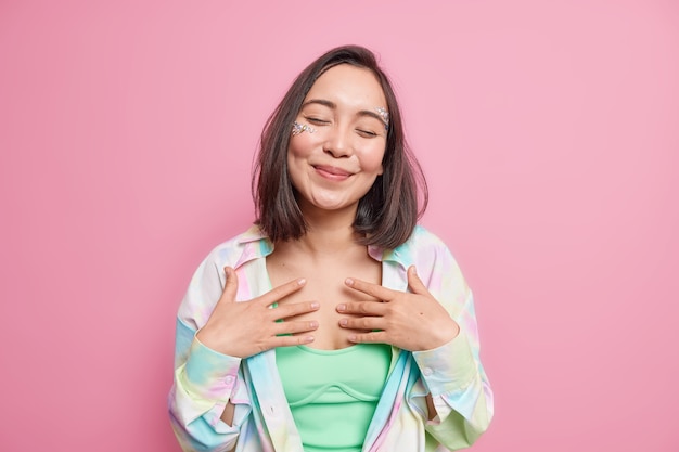 Tender cheerful Asian woman keeps eyes closed hands on chest recalls nice memories smiles tenderly pictures nice romantic scene in mind dressed casually isolated over pink  wall.