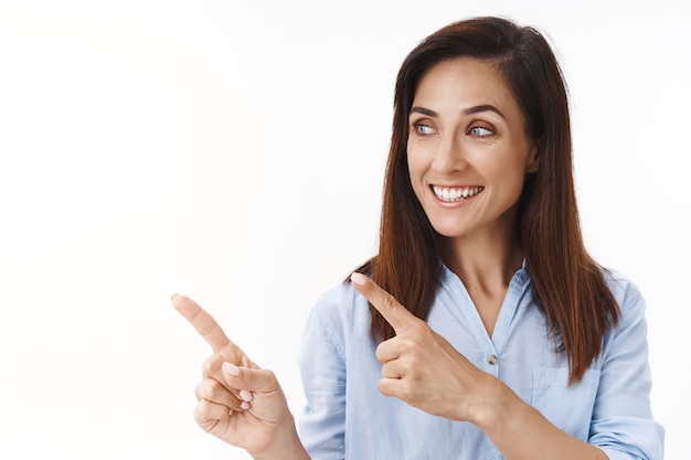 Tender cheerful middle-aged woman with perfect clean skin no makeup, pointing turn left copyspace, smiling enthusiastic, observe promo with interest and joy, grinning delighted, white wall