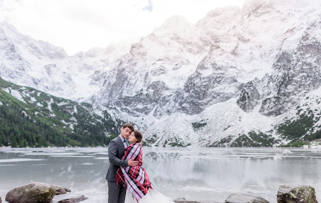 Free photo tender couple covered with blanket in love freezing in winter mountains in front of frozen lake