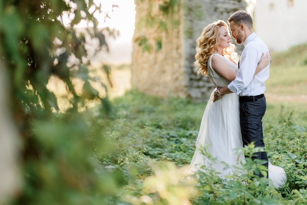 Tender couple is almost kissing outdoors on the warm sunny day near the stone building surrounded with green grass