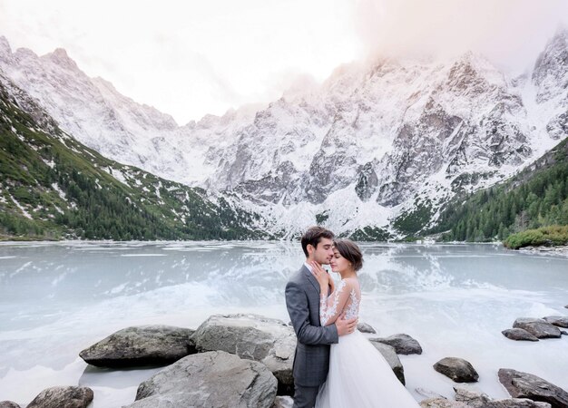 凍った湖と雪に覆われた高山の前で結婚式の衣装に身を包んだ愛の優しいカップルが抱いています。