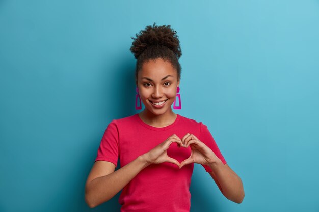 Tender dark skinned young woman makes heart gesture, shows love, affection, passion, wears casual rosy t shirt and earrings, isolated on blue wall. Body language, romance, feelings concept