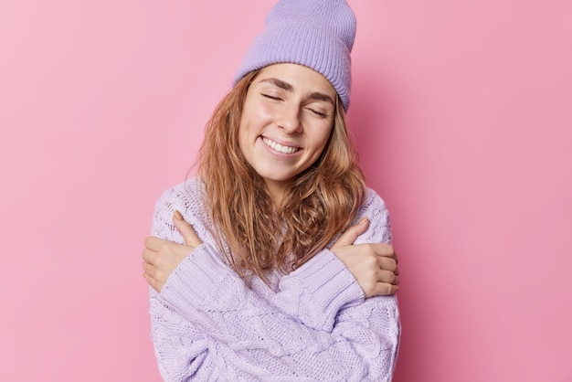 La tenera e delicata giovane donna sorridente si abbraccia con amore gode di comfort nel maglione lavorato a maglia tiene gli occhi chiusi indossa il cappello pone su sfondo rosa ha bei ricordi romantici si sente morbido