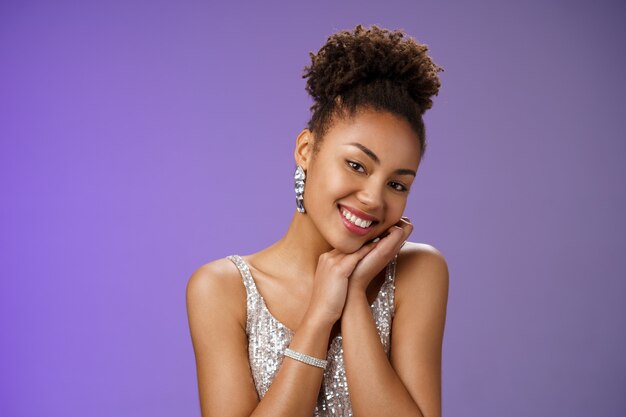 Tender elegant wealthy young african american rich woman in brilliants silver stylish shiny dress lean head shoulder smiling flirty romantic gaze camera standing amused blue background.