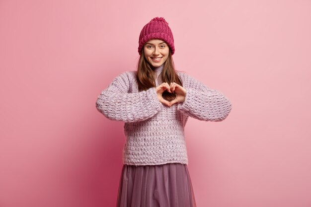 Free photo tender feminine girl shapes heart with hands over chest, expresses love and sympathy, wears pink headgear