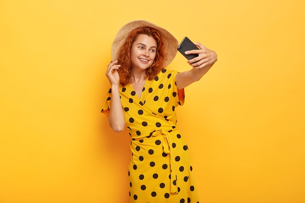 Tender feminine girl with ginger hair, takes selfie on smartphone, smiles broadly