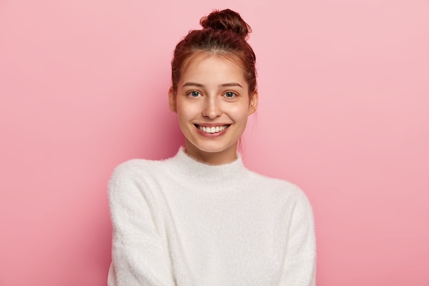 Tender feminine woman with blue eyes, smiles pleasantly, has toothy smile, wears white comfortable sweater, looks directly at camera, isolated on pink background