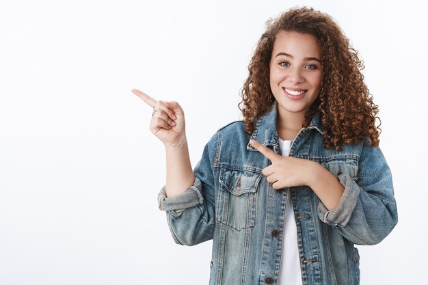 Tender feminine young female student curly hairstyle pointing left index finger smiling friendly asking which place dorm standing cheerful white wall