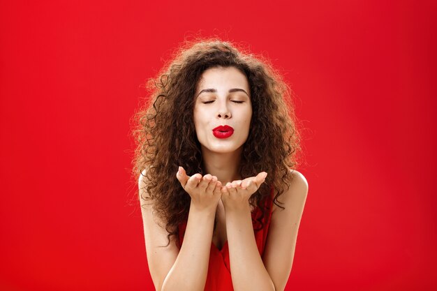 Tender and gentle stylish caucasian girl with curly hairstyle and red lipstick bending towards camera with slight smile closed eyes and palms near folded lips blowing kiss at camera romantically.