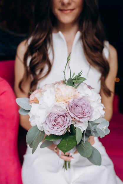 Tender and lovely bride sits with rich wedding bouquet on a pink couch in the cafe