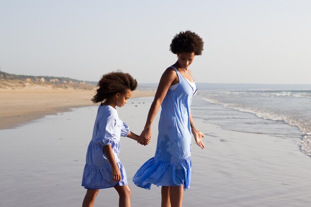 부드러운 엄마와 딸 휴가입니다. 아름다운 드레스를 입은 아프리카계 미국인 가족은 해변을 걷고, 좋은 시간을 보내고, 손을 잡고 있습니다. 가족, 여행, 부모 개념