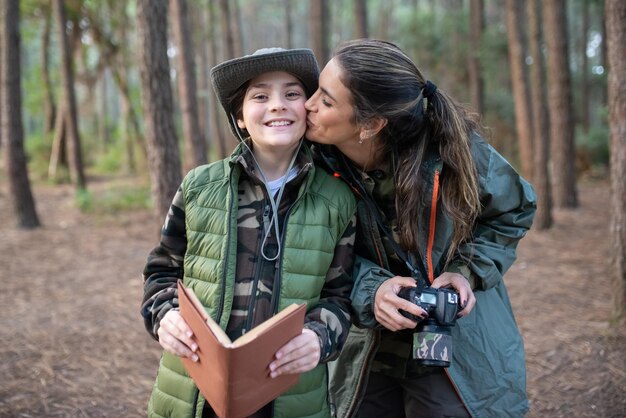 森の中でカメラを持つ息子と優しい母親。頬に笑みを浮かべて少年にキスをするスポーツ服の女性モデル。趣味、写真のコンセプト