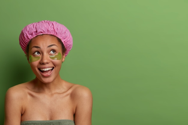 Tender positive dark skinned woman with collagen eye pads, concentrated above, smiles positively, has bare shoulders, wears bath cap, has pure clean skin. Body care routine. Green wall