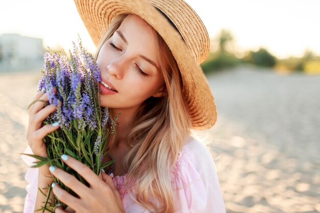 花の花束と海の近くの太陽が降り注ぐビーチでポーズをとる麦わら帽子の優しいきれいな女性。肖像画をクローズアップ。