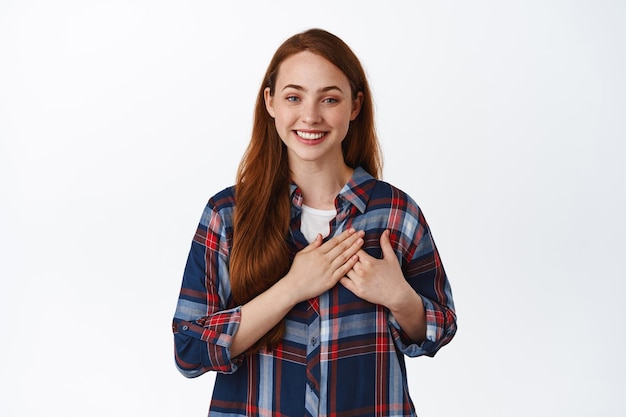 Free photo tender redhead girl with lovely smile, hold hands on heart, being flattered, grateful or delighted, say thank you, appreciate efforts, white background