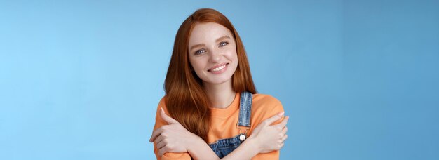 Free photo tender silly redhead girl standing blue background smiling joyfully hugging arms crossed body feel