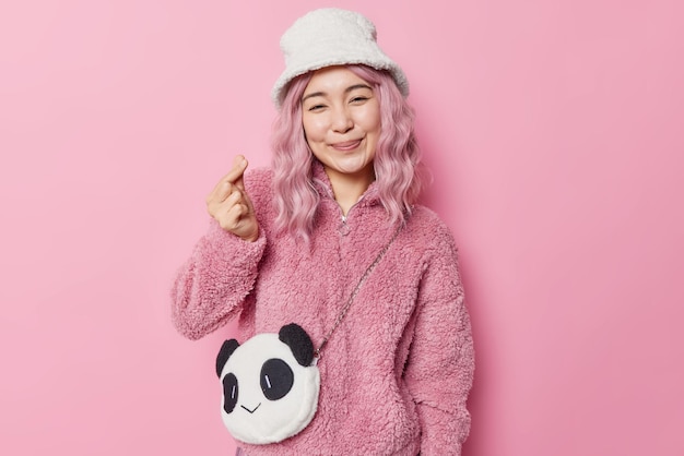 バラ色の髪の優しい笑顔のアジアの女性は、サインのような韓国語が愛の形を表現していることを示していますミニハートは毛皮のコートを着ており、パナマはピンクの背景の上に分離されたバッグのようなパンダを持っています。ボディーランゲージの概念