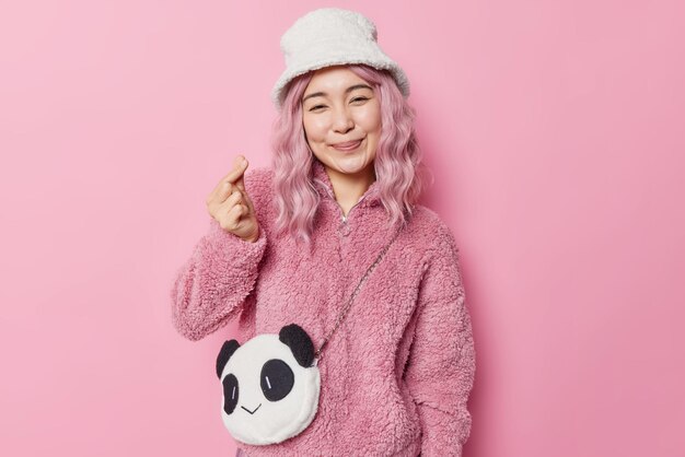 Tender smiling Asian woman with rosy hair shows korean like sign expresses love shapes mini heart wears fur coat and panama has panda like bag isolated over pink background. Body language concept