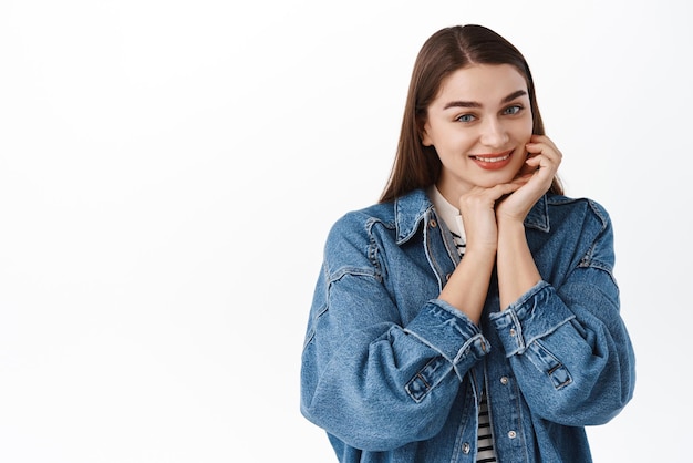 Tender smiling woman gently touching her face holds hands near skin and smiles flirty at camera making innocent angelic expression standing over white background Copy space