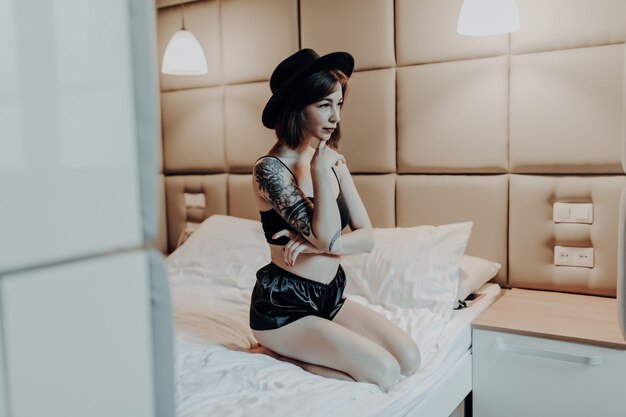 Нежная крошечная женщина в модном черном нижнем белье и широкой шляпе сидит утром на кровати