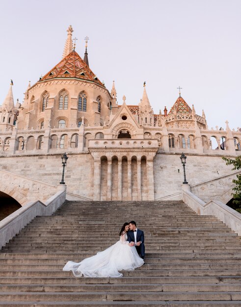 暖かい夜に古い石造りの歴史的な建物の階段に優しい結婚式のカップルが座っています。