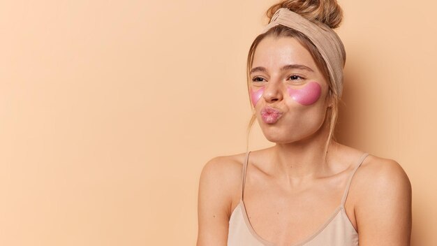 Tender woman with healthy smooth skin keeps lips rounded wears hydrogel patches under eyes to reduce fine lines undergoes beauty treatments wears headband poses indoor against brown background