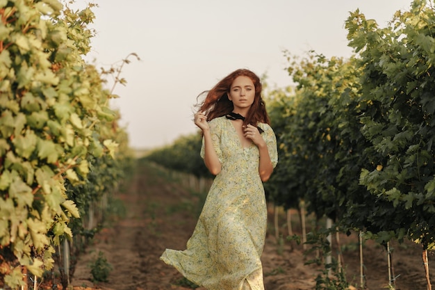 Tender woman with red wavy hairstyle and black bandage on neck in long stylish summer dress looking at front on a vineyard