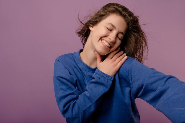 Tender young caucasian brunette girl in blue hoodie smiles with her eyes closed on purple background Emotions and states of mind concept