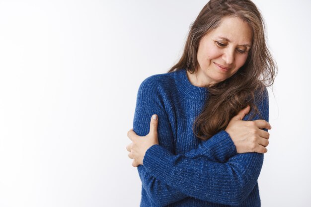 Tenderness and family concept. Gentle and tender lovely middle-aged woman in sweater hugging herself with hands crossed on arms looking down shy and cute, smiling against white wall