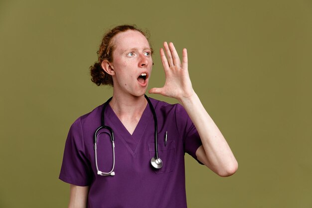 Tense calling someone young male doctor wearing uniform with stethoscope isolated on green background