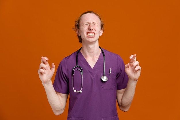 напряженные скрещенные пальцы молодой врач-мужчина в униформе со стетоскопом на оранжевом фоне