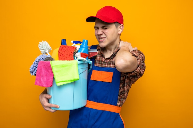 Tense grabbed aching neck young cleaning guy wearing uniform and cap holding bucket of cleaning tools