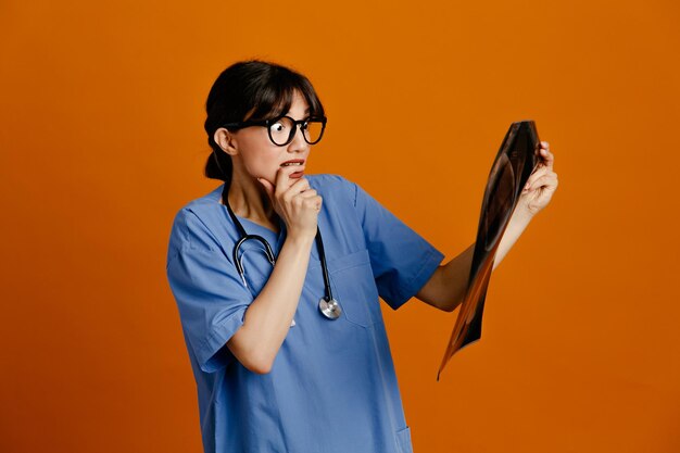 オレンジ色の背景に分離された制服 fith 聴診器を身に着けている x 線若い女性医師を保持している緊張したあご